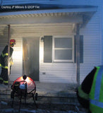 All-Terrain Ventry Fan Model 24GX160 with optional Top Light LED and Large Pneumatic Tires shown with the light on, lighting the entry way to a house during a fire department's evening training. The fan is placed on the ground in front of the deck with the three independently adjustable legs partically extended, allowing the fan to be raised above the deck, leaving the entryway clear.