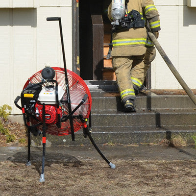24-inch VENTRY Fan Model 24GX160 with optional Medium Flat-Free Wheels & Skids and Top Light LED shown during a fire department's PPV training. The fan is shown set off to the side with the three independently adjustable legs extended at different lengths, allowing the fan to be aimed over the stairs and into the front door. This provides clear access for the firefighter carrying a hose into the door/house.